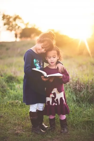Two girls reading a book together in a field at sunset.